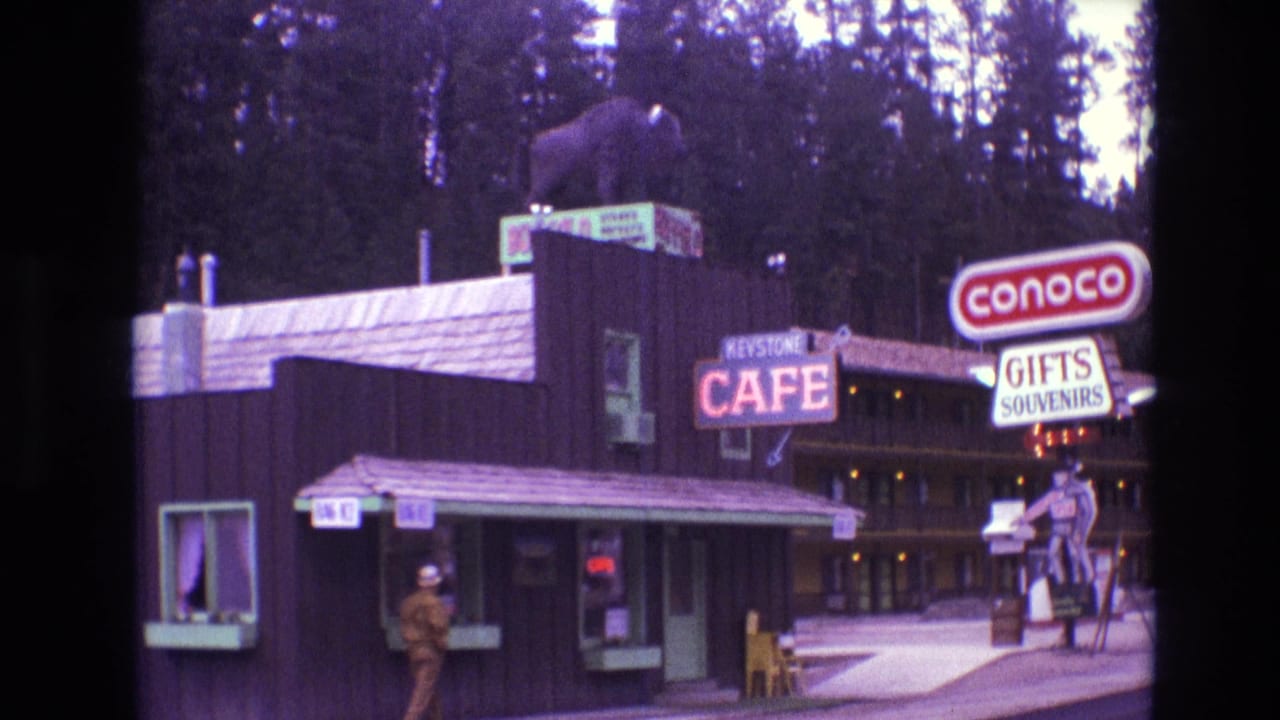 1974 South Dakota 8mm frame showing the Keystone Cafe exterior, a roof-mounted bear statue, a Conoco sign, Gifts Souvenirs signage, and an adult man near the service window.