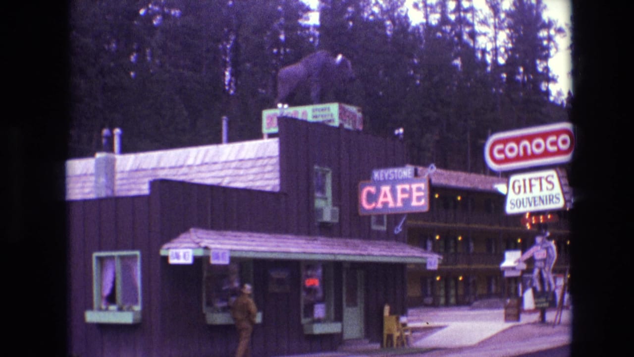 Roadside cafe building with roof bear statue, Conoco sign, Gifts Souvenirs sign, and man by the counter.