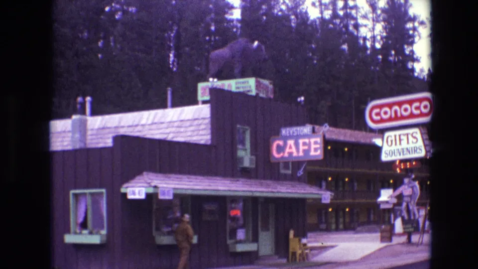 1974 South Dakota frame of the Keystone Cafe roadside stop with a bear statue above the roofline, Conoco branding, souvenir signage, and an adult visitor standing near the front counter.