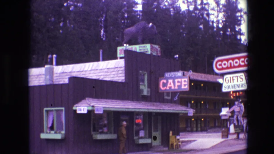 1974 South Dakota 8mm frame showing the Keystone Cafe from a slightly shifted angle, with service windows, roof bear statue, cafe lettering, partial Conoco sign, and forest behind the buildings.