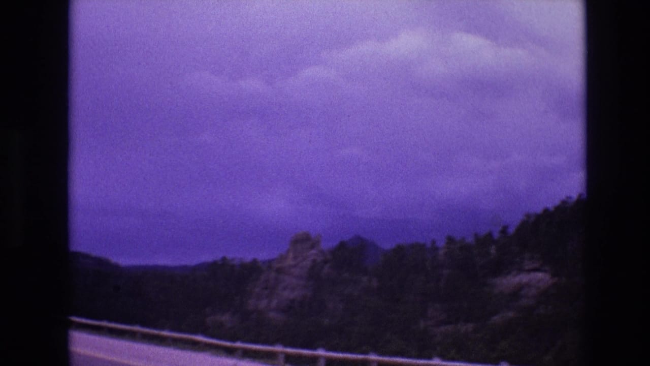 Cloudy purple-tinted sky over dark wooded hills and rocky terrain beside a roadside railing.