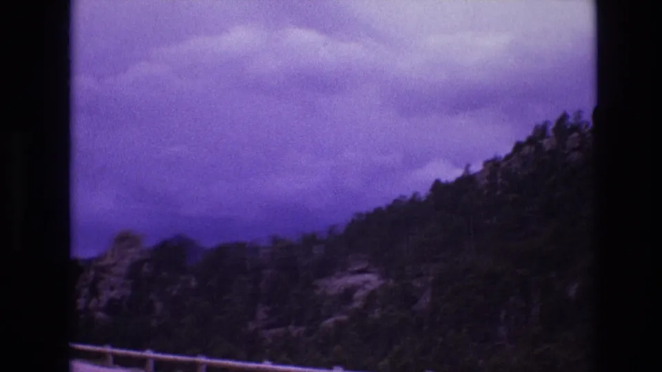 1974 South Dakota 8mm frame with a pine-covered hillside, rocky slope, roadside railing, and layered clouds rendered with strong home-movie color shift.