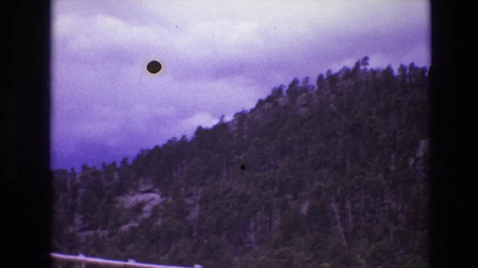 1974 South Dakota 8mm frame showing a wooded slope with exposed rock beneath a cloudy sky, with visible dust specks and soft film texture from the scan.