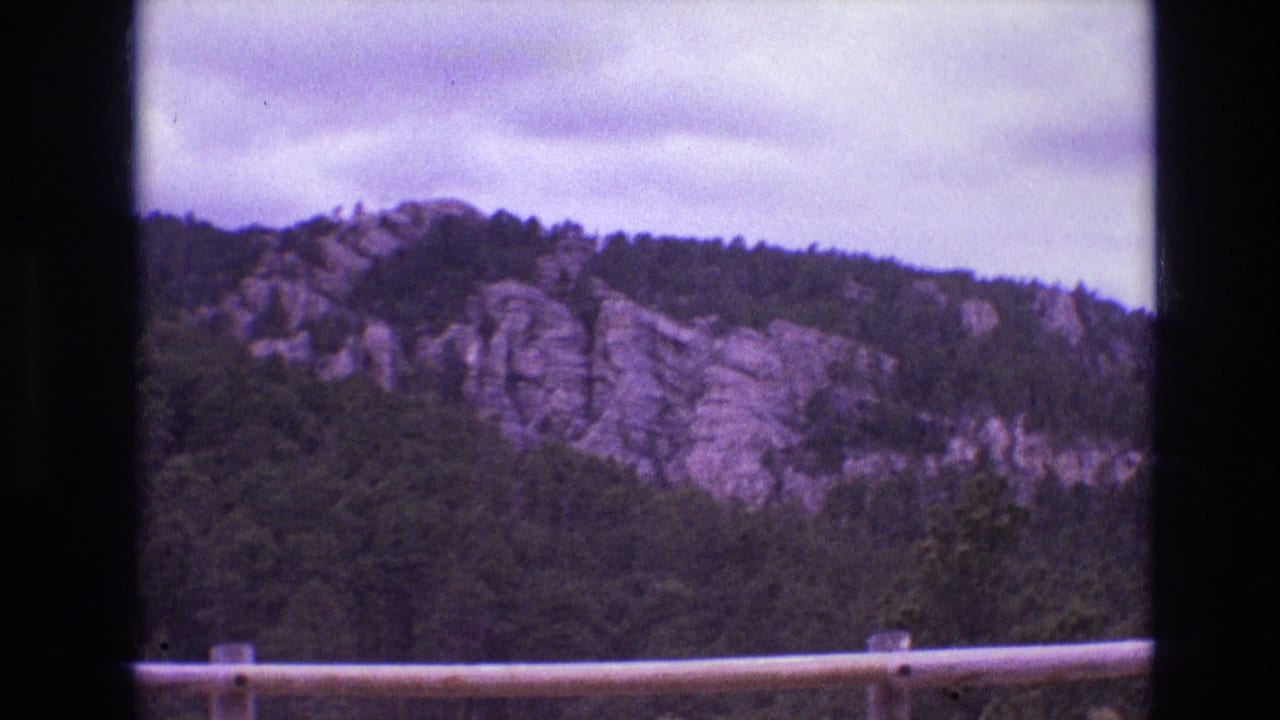 1974 South Dakota 8mm frame showing a distant forested ridge with pale rock formations, cloudy sky, and a roadside guardrail in the foreground.
