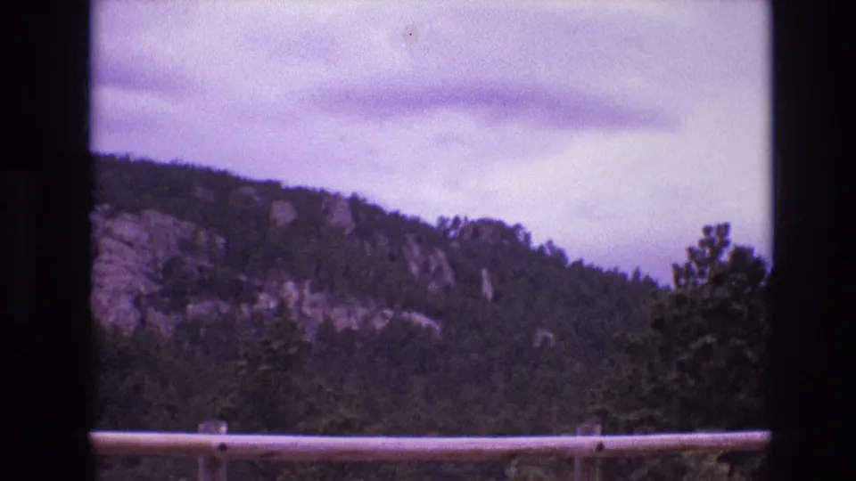 1974 South Dakota 8mm frame showing the ridgeline shifted lower in view, with forest, exposed rock, cloudy sky, and roadside guardrail visible.