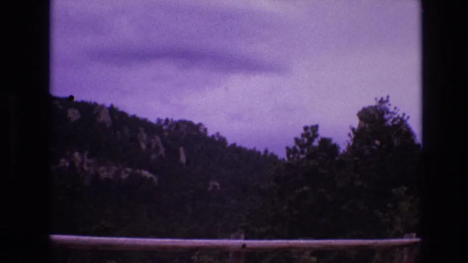 1974 South Dakota 8mm frame with a darker exposure of the wooded ridge, rocky patches, cloudy sky, and foreground roadside guardrail.