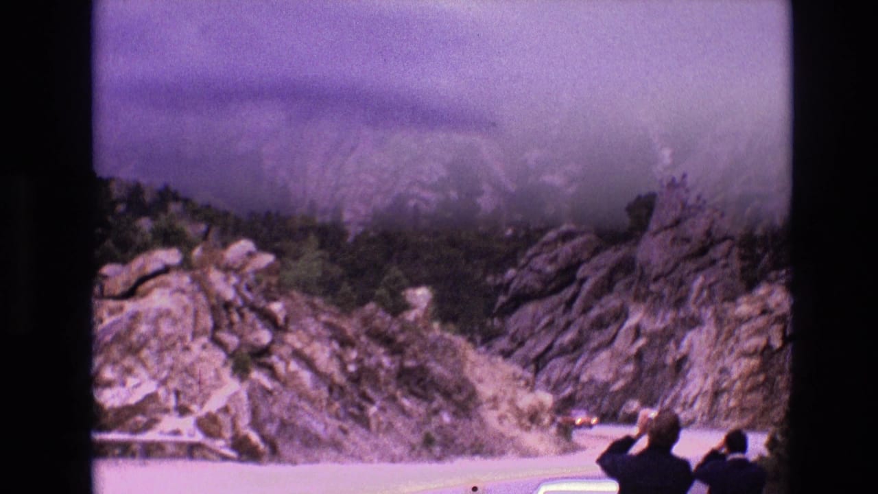 1974 South Dakota 8mm frame showing rocky bluffs, evergreen brush, a curving road, and two roadside visitors looking upward near a light-colored car.