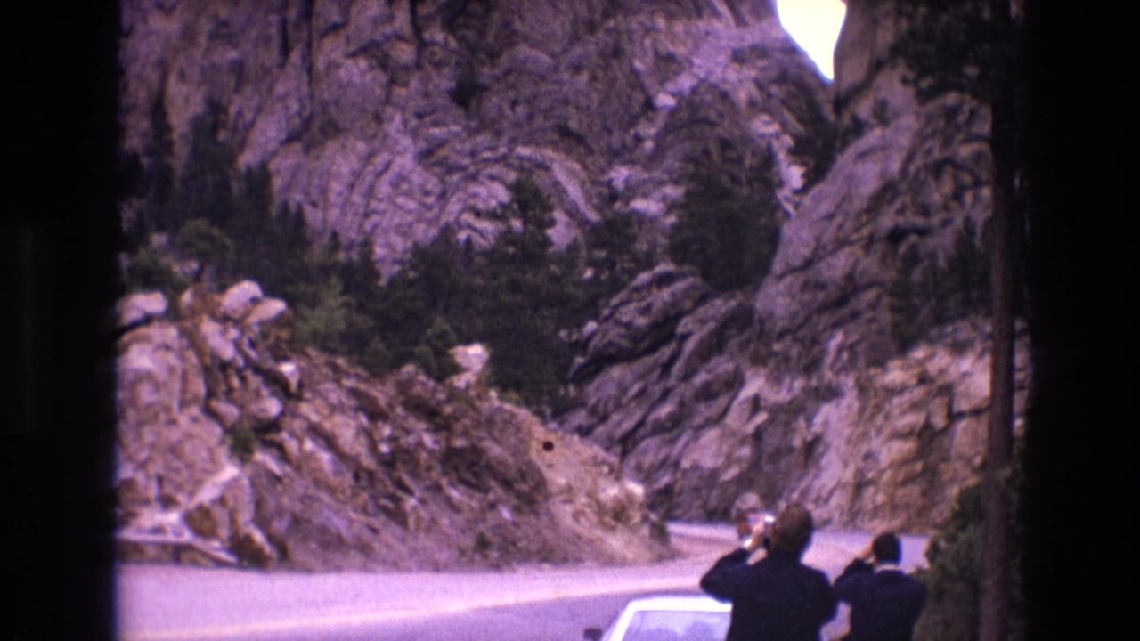Two people stand beside a light-colored car on a road below steep rock formations.
