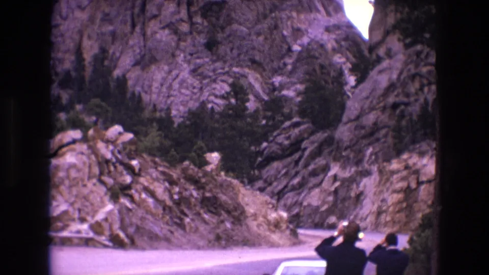 1974 South Dakota 8mm frame of a winding paved road through rocky bluffs, with two visitors near a car and scrubby evergreen growth along the slope.