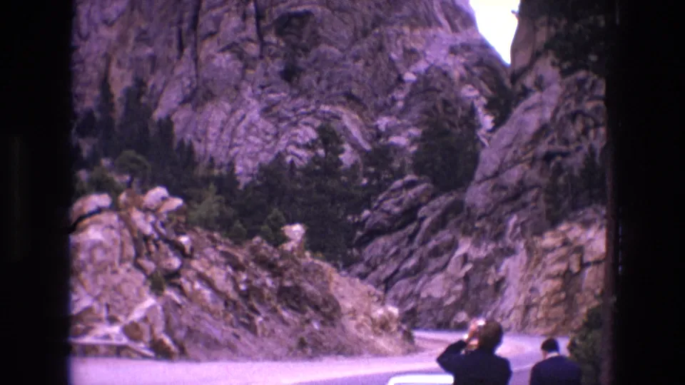 1974 South Dakota home-movie frame showing two roadside visitors near a parked light-colored car, a narrow road bend, dense rock faces, and dark evergreen brush.