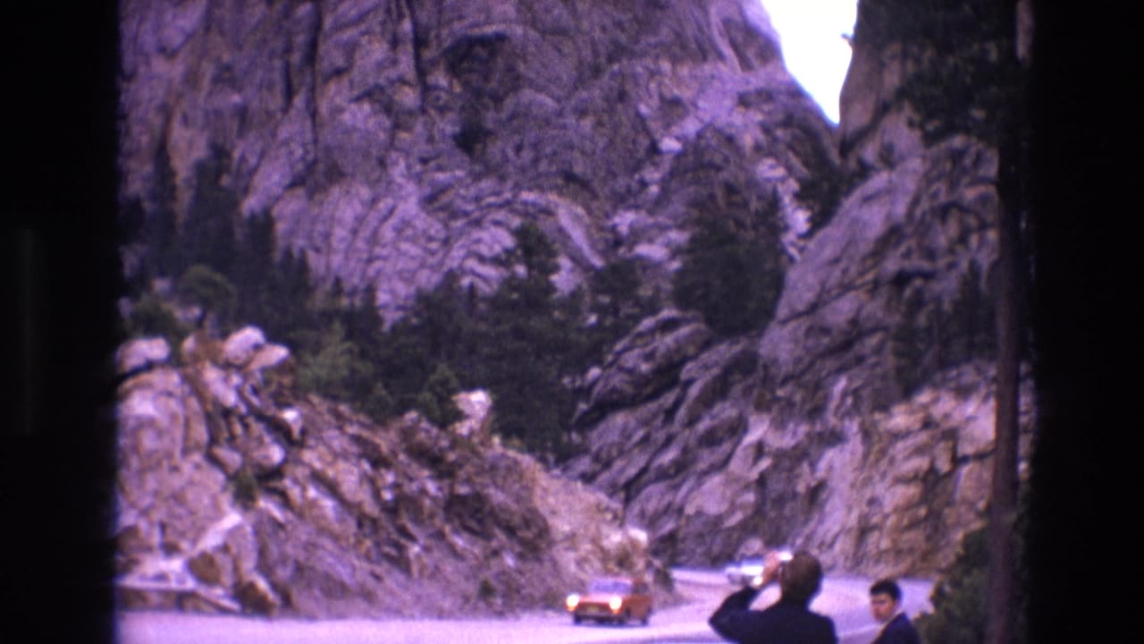 Orange car rounds a curve between rocky cliffs as two men stand at the roadside.