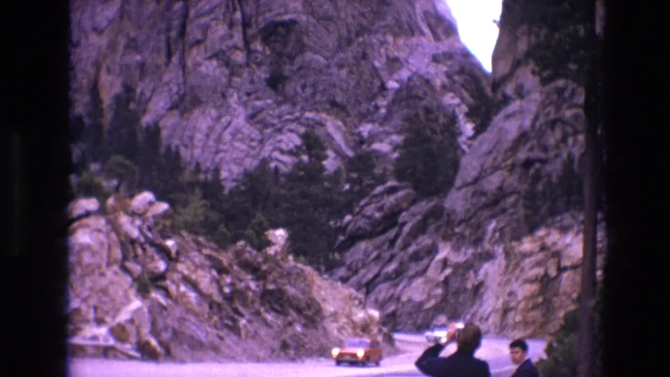 1974 South Dakota 8mm frame showing an orange car navigating a curved mountain road while two adult men stand near the edge of the road.