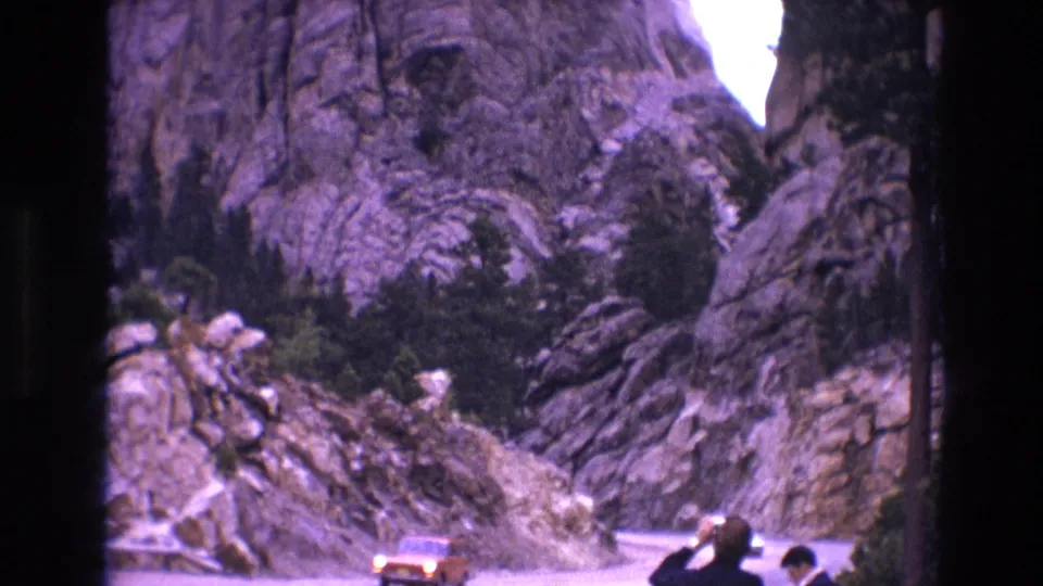 1974 South Dakota 8mm frame showing an orange car on a winding paved road below rugged cliffs and scattered evergreens.