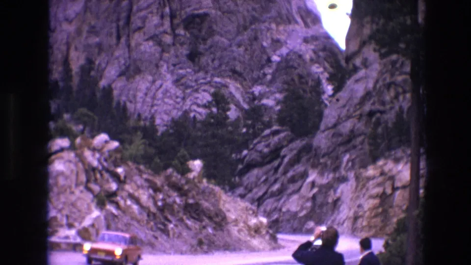 1974 South Dakota 8mm frame showing two adult men at the roadside as an orange car passes through a curve beneath rocky cliffs.