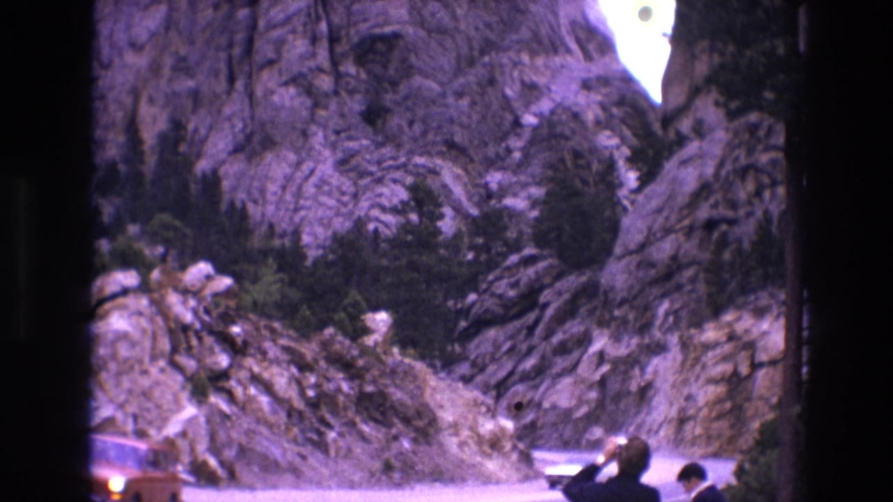 1974 South Dakota 8mm frame showing a classic car on a road below steep rock formations, with roadside visitors partly visible in the foreground.