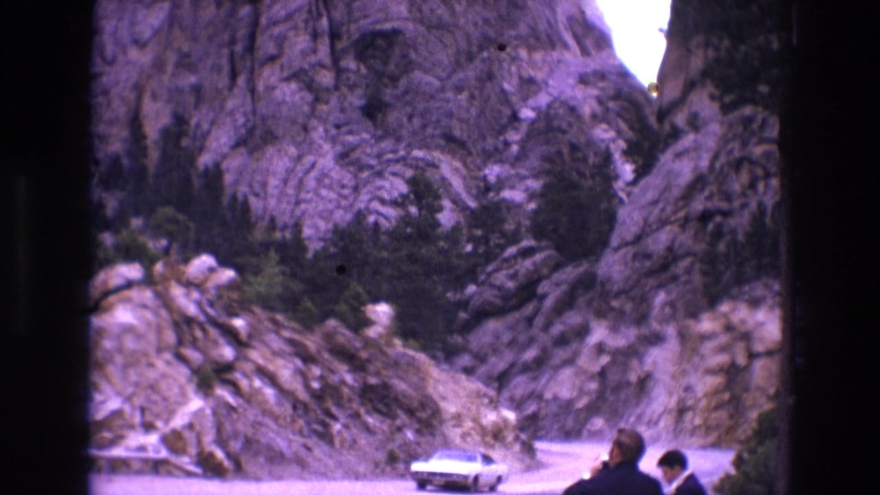 1974 South Dakota home-movie frame of a light-colored classic car traveling through a rocky pass, with two roadside figures partly visible at frame edge.