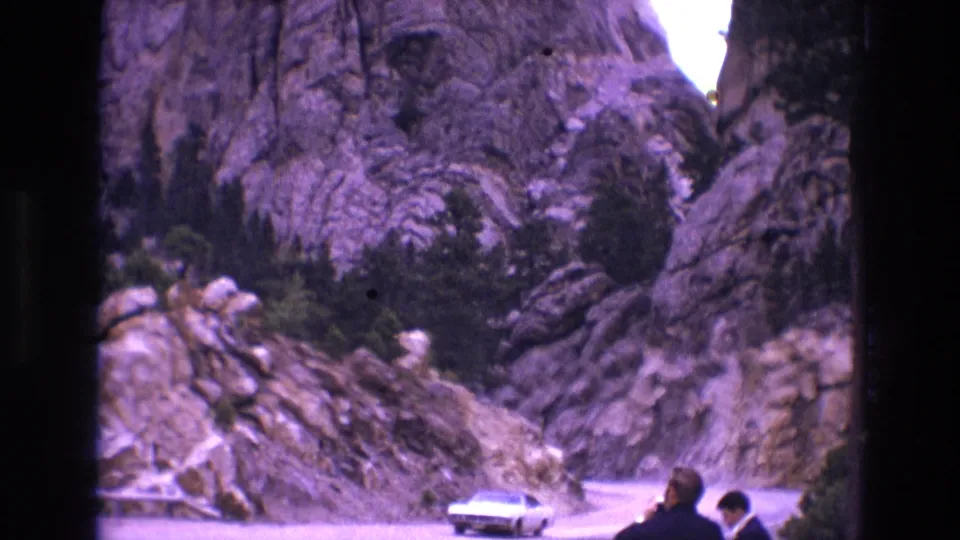 1974 South Dakota home-movie frame of a light-colored classic car traveling through a rocky pass, with two roadside figures partly visible at frame edge.