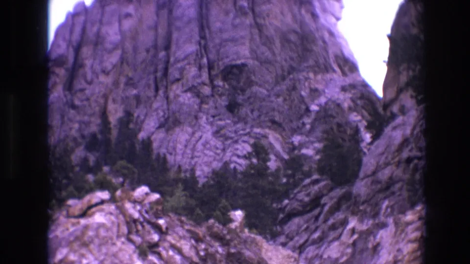 1974 South Dakota 8mm frame showing rugged rocks and evergreens beside a mountain-pass road, with the car no longer prominent in view.