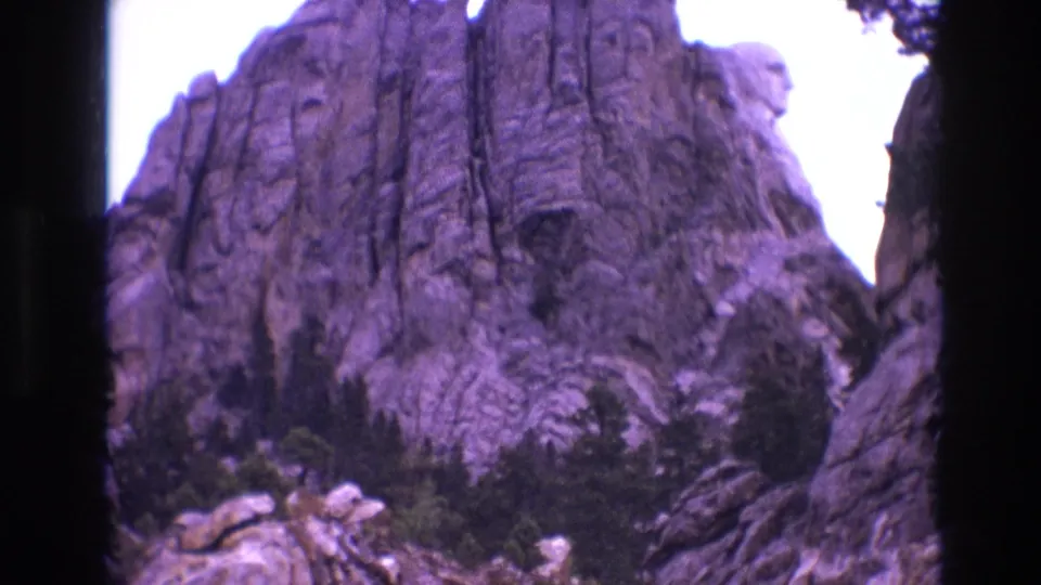 1974 South Dakota home-movie frame looking up at steep rock formations and trees in a mountain pass, framed by visible 8mm gate edges.