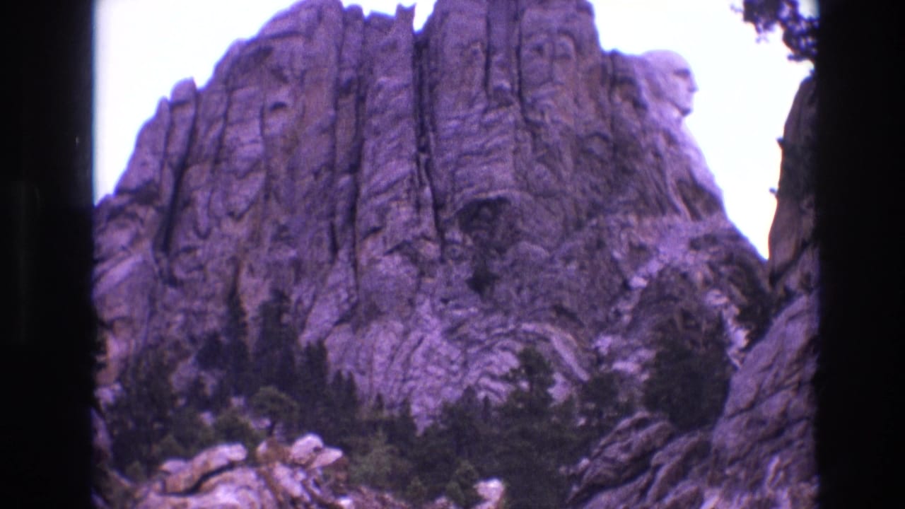 1974 8mm frame showing Mount Rushmore in South Dakota from below, with granite presidential faces above evergreen trees and a pale sky.
