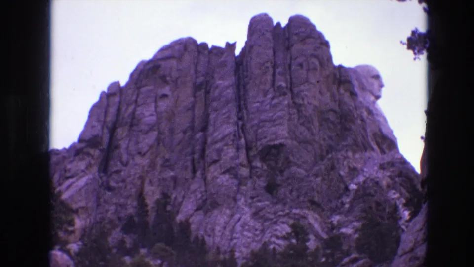 1974 8mm travel frame of Mount Rushmore from a low viewpoint, showing the granite cliff face, sculpted profiles, trees, and purple film color cast.