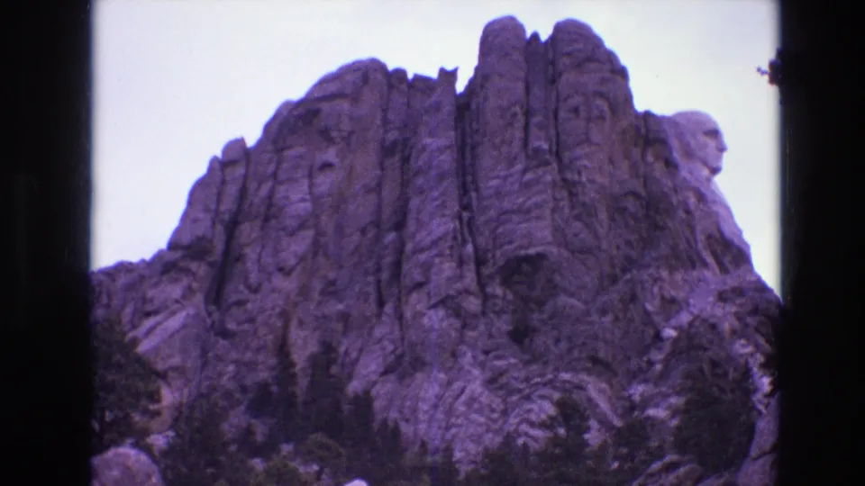 South Dakota 1974 home-movie frame showing Mount Rushmore from below, with presidential carvings in granite above a dark evergreen line.