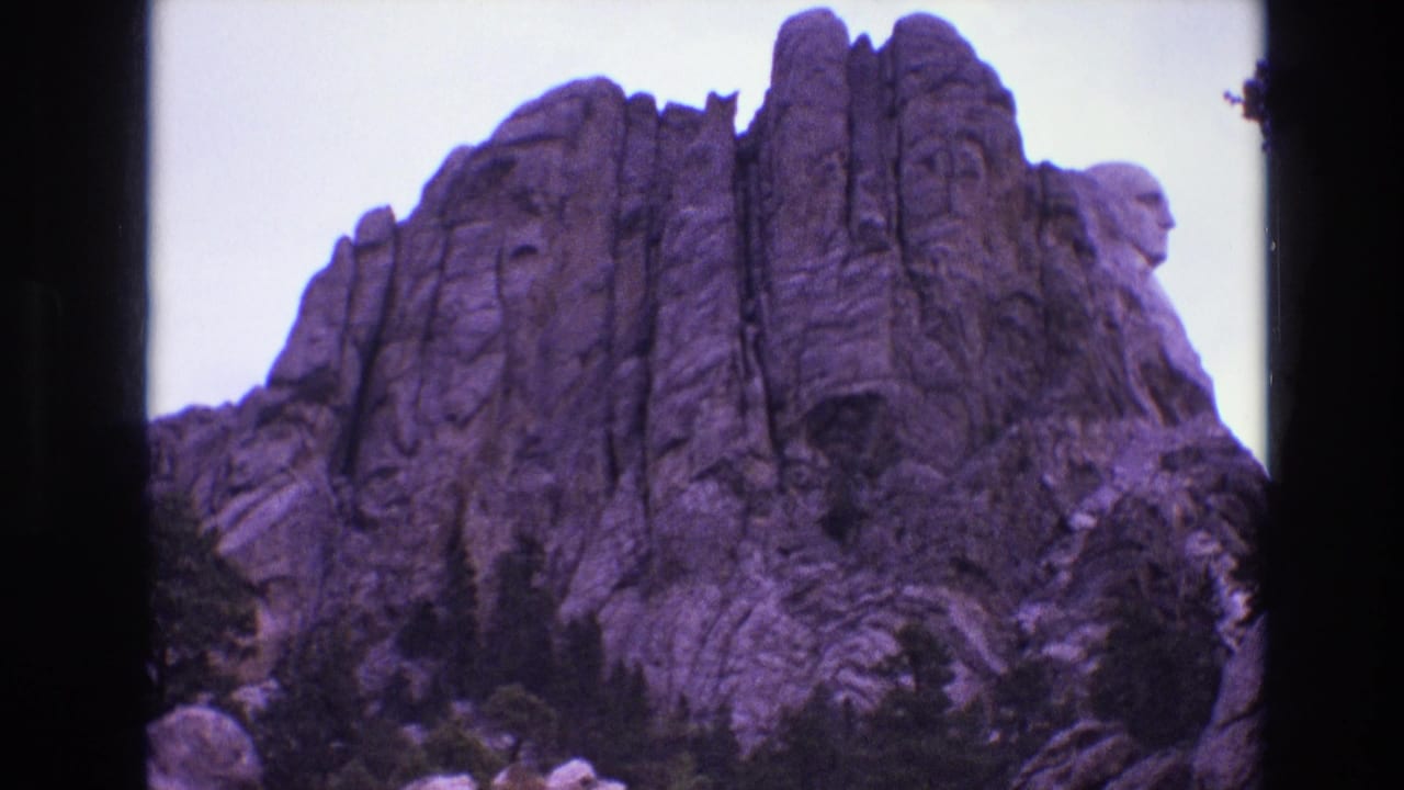 1974 8mm frame of Mount Rushmore in South Dakota, showing the carved presidential heads above rugged granite and evergreen trees with a purple film cast.