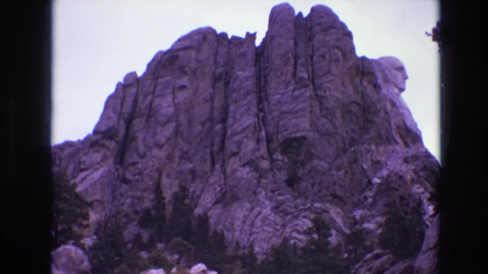 1974 8mm frame of Mount Rushmore in South Dakota, showing the carved presidential heads above rugged granite and evergreen trees with a purple film cast.