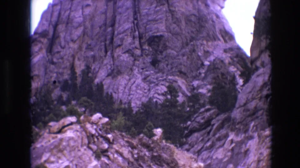 1974 8mm view of Mount Rushmore from a lower roadside vantage, with carved heads partly high in frame, rugged stone, trees, and aged-film color shift.