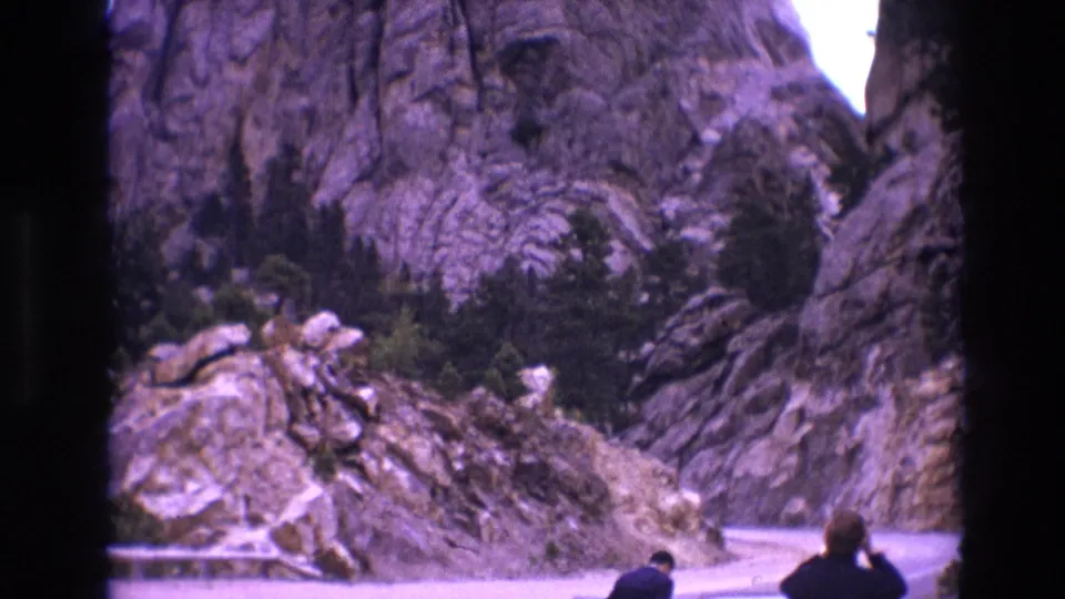 1974 South Dakota frame of Mount Rushmore and surrounding rocks above a curving road, with visitors at the lower right and one person appearing to look through binoculars.