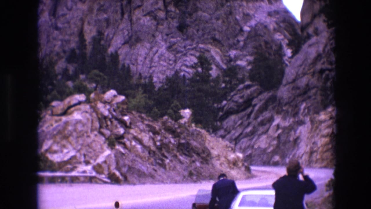 1974 South Dakota 8mm frame showing roadside visitors beside a white car and another vehicle on a curving road near rugged rock formations.