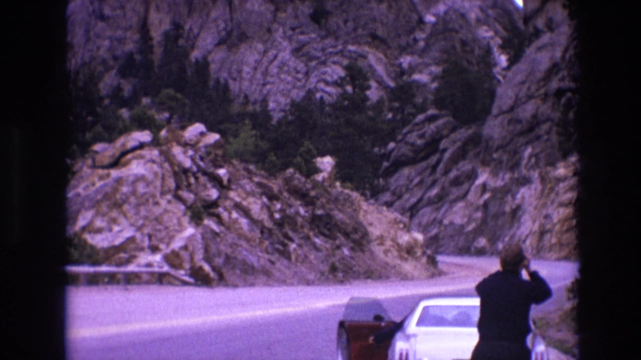 A person stands near a white car on a curving road bordered by rock formations.