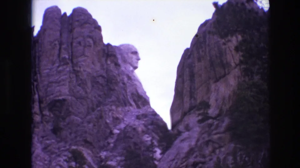 1974 South Dakota 8mm frame showing the carved faces of Mount Rushmore partially framed by dark rock walls and pale sky.