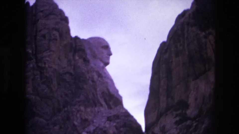 1974 South Dakota home-movie frame with Mount Rushmore visible in the distance, partially enclosed by rugged foreground cliffs.