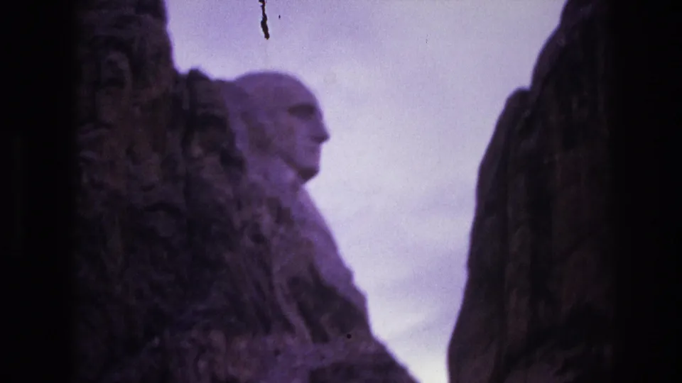 1974 8mm frame of Mount Rushmore with a carved presidential profile visible behind dark foreground rock and a pale, color-shifted sky.