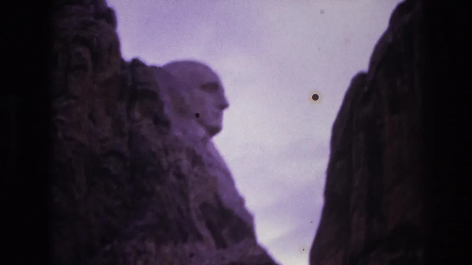 1974 8mm frame of Mount Rushmore in South Dakota, with a carved profile between dark rocks and small circular film or optical marks visible against the sky.