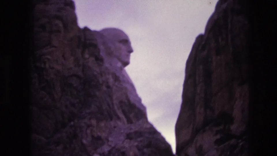 1974 Mount Rushmore home-movie frame with Washington's carved side profile seen beyond shadowed granite columns and a pale purple-tinted sky.