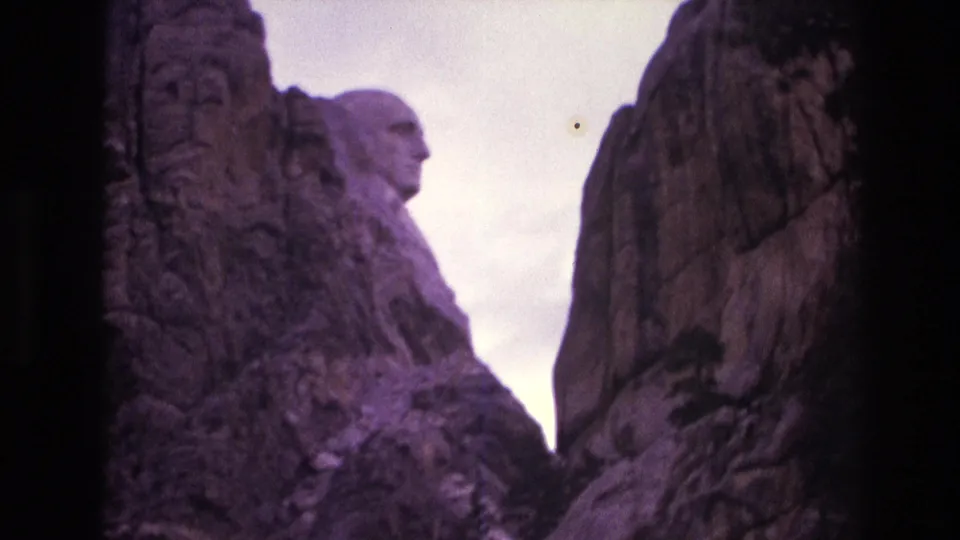 Low-angle 1974 South Dakota travel frame showing Mount Rushmore's granite surfaces with Washington's profile partially visible behind a darker rock wall.