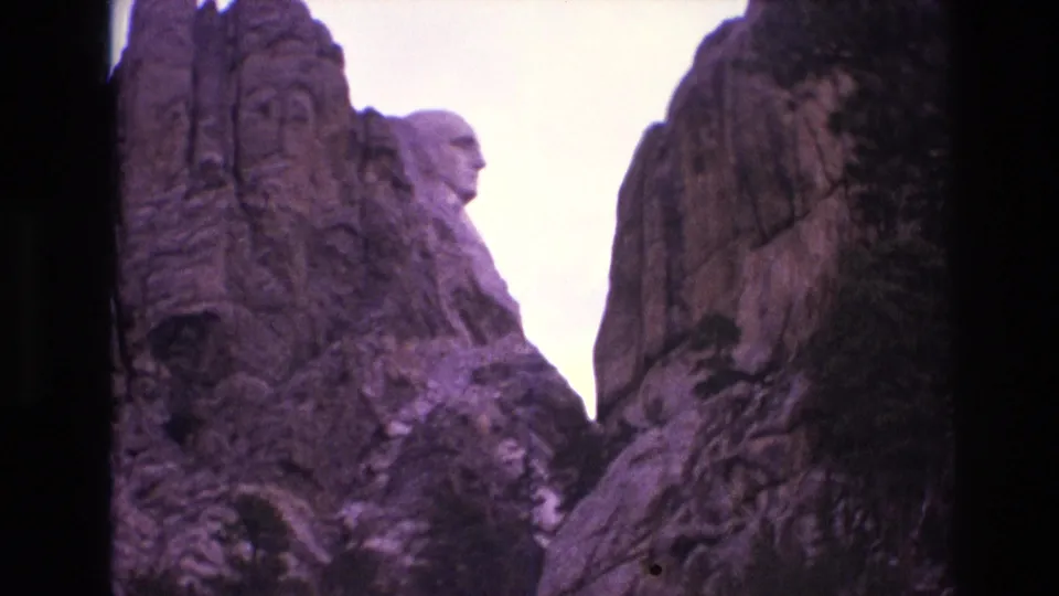 1974 8mm frame from Mount Rushmore showing a closer side view of sculpted granite and George Washington's profile against a bright, purple-tinted sky.