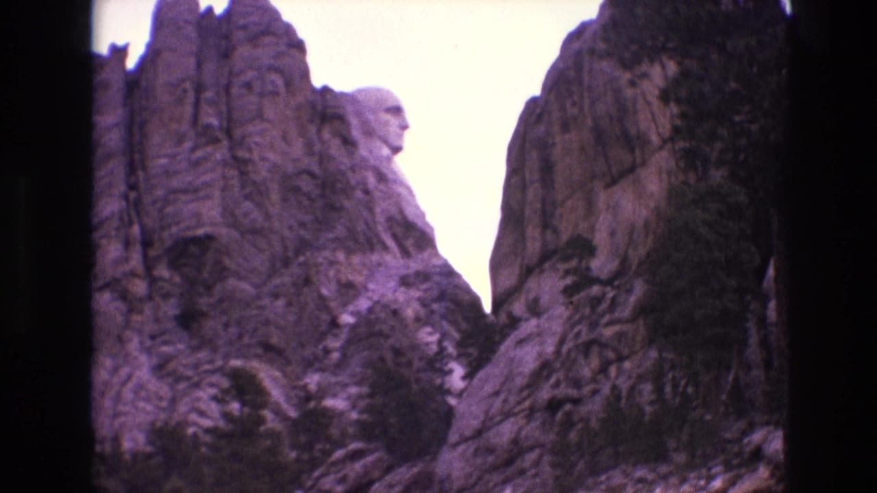 1974 8mm home-movie frame looking up at Mount Rushmore in South Dakota, with carved faces set above granite outcrops and dark film borders.