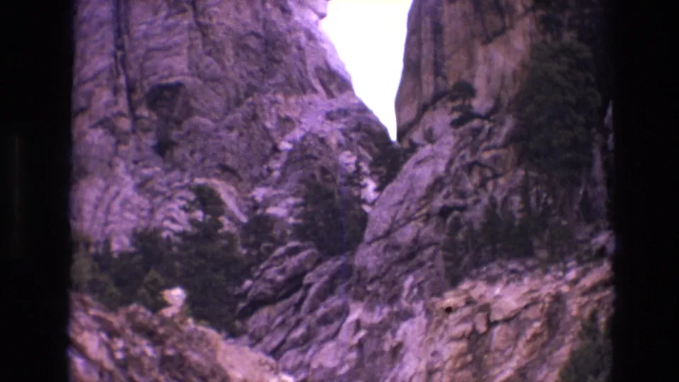 1974 home-movie frame from below Mount Rushmore, showing the monument partly obscured by granite outcrops, trees, and aged-film edge shadows.