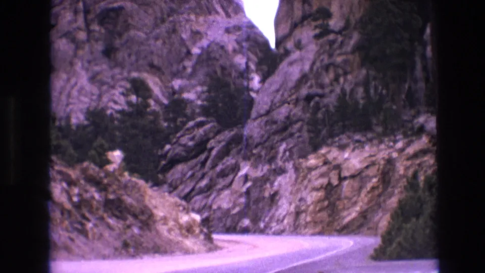 The camera has panned down to a winding paved road below the Mount Rushmore rock formations, with granite slopes and trees framing the 8mm travel view.