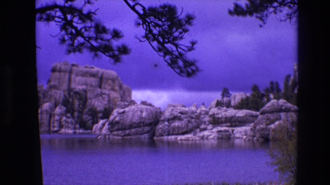 1974 South Dakota 8mm frame of a calm lake with rounded granite-like rock formations and pine branches silhouetted near the top edge.