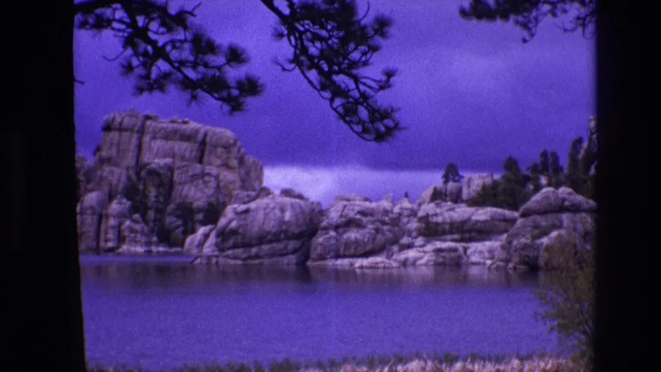 1974 South Dakota 8mm travel frame showing still lake water, massive rounded rocks, overhanging pine branches, and low vegetation along the foreground edge.