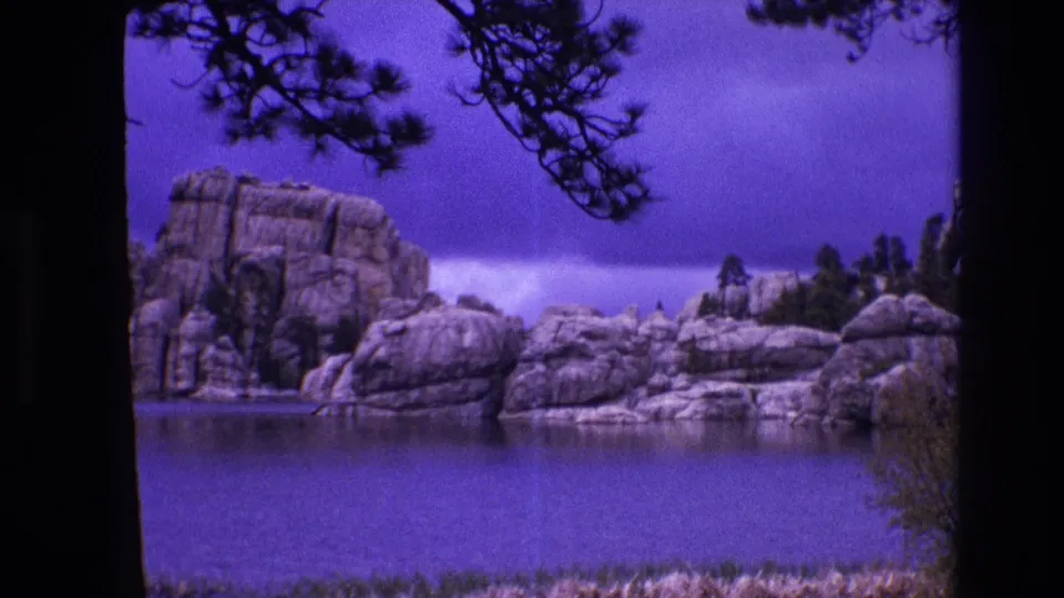 1974 South Dakota 8mm frame of calm water facing rounded lakeside rock formations, with pine branches across the upper frame and visible film scan borders.