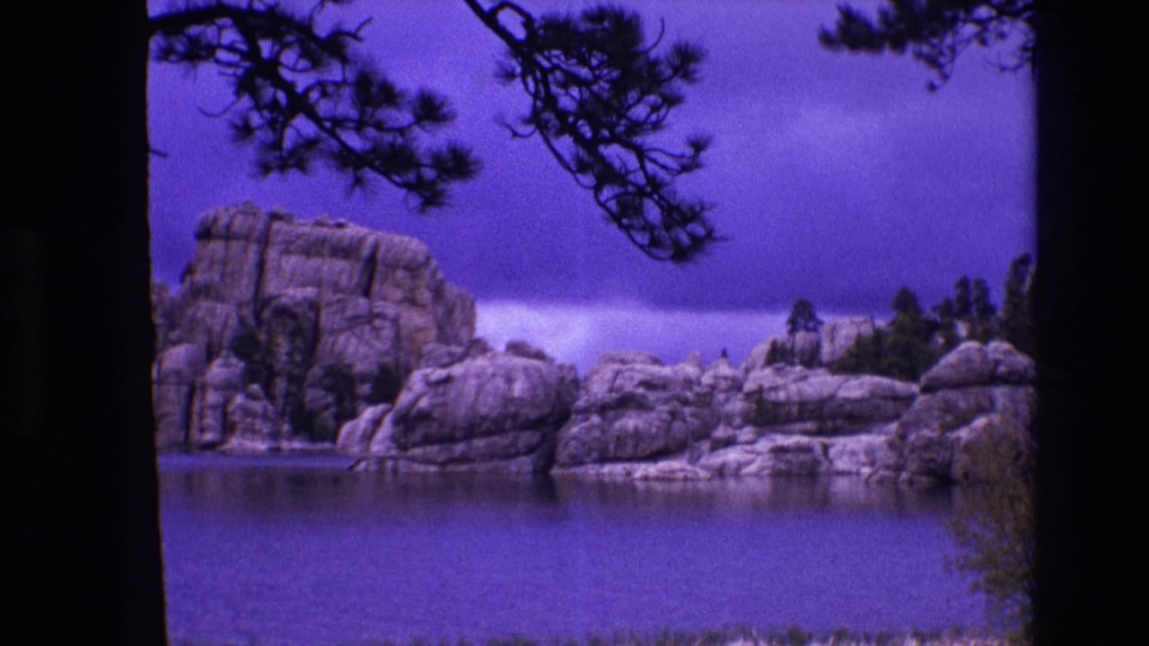 1974 South Dakota 8mm frame showing a calm lake, large rocky formations, pine branches, and dark tree trunks at the edge of the shot.