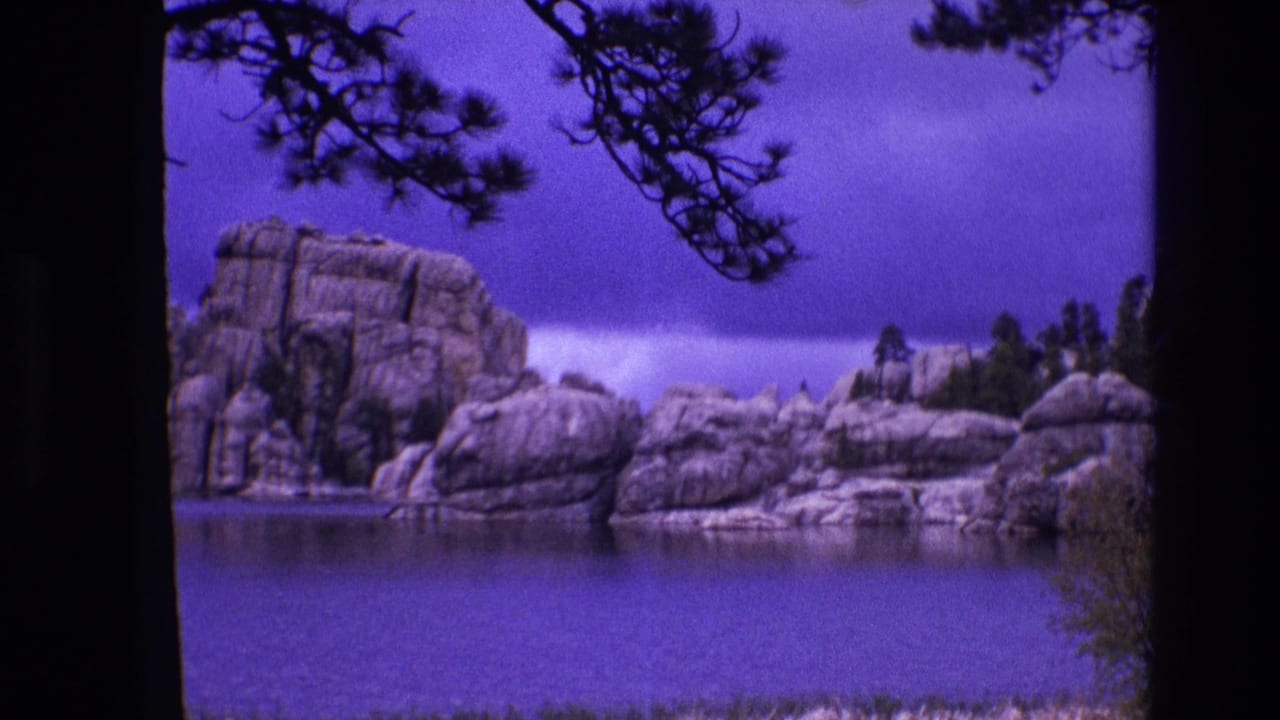 1974 South Dakota 8mm frame with a still lake, pale rock outcrops, pine branches overhead, and a strong purple cast from aged color film.