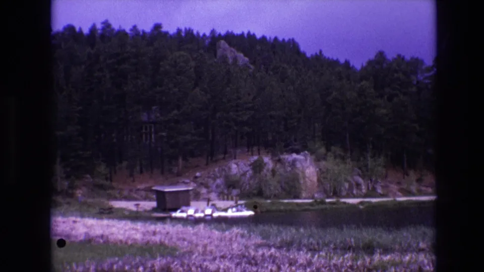 1974 South Dakota 8mm frame showing a forested shoreline, small lakeside structure, dock, boat, reeds, and rocky rise near the water.