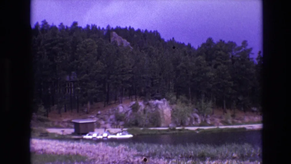 1974 South Dakota 8mm frame of a wooded lakeshore with a docked boat, small structure, rocky bank, reeds, and purple-shifted sky.
