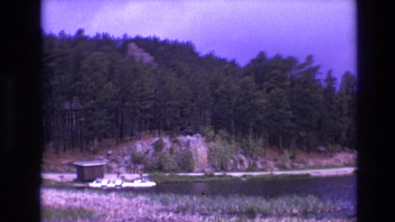 1974 South Dakota 8mm frame showing a calm lake bordered by pine trees and rocky shoreline, with a small boathouse, dock, and white boat near the water.